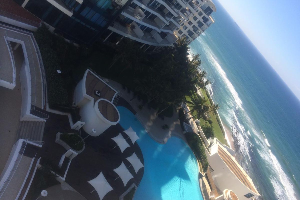 An elevated aerial view of the apartment complex pool area, featuring landscaped grounds and direct access to the coastline.