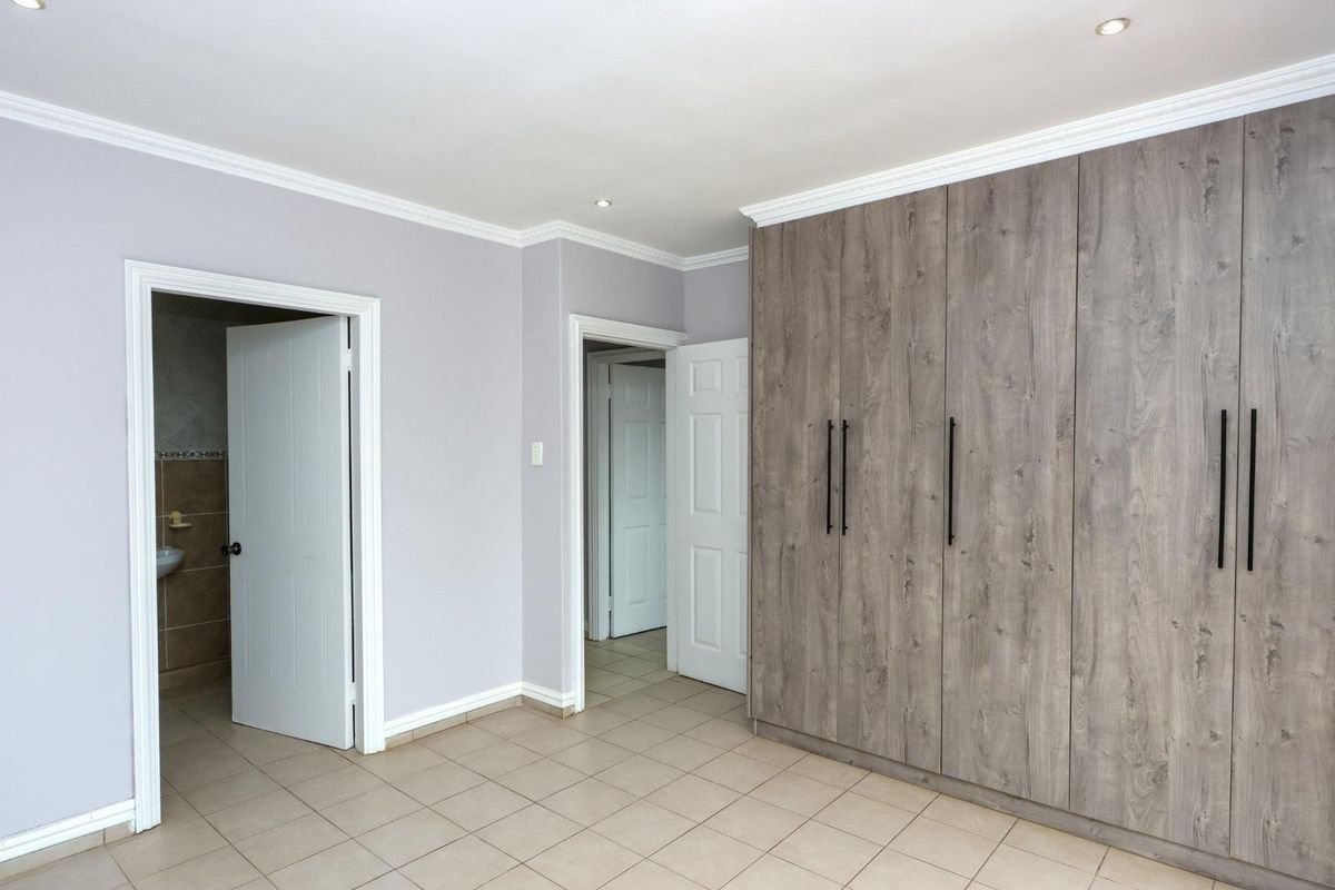 A spacious bedroom featuring modern wood-finish built-in cupboards, neutral tiled flooring, and recessed downlighting, with direct access to an en-suite bathroom.