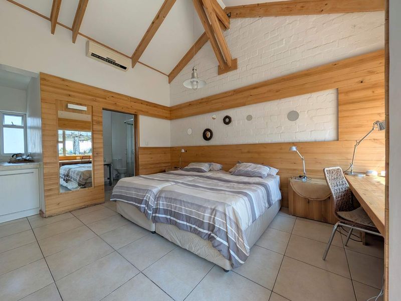 Spacious bedroom featuring exposed timber roof trusses, timber wall paneling, painted brickwork, tiled flooring, and an integrated desk area with en-suite bathroom access.