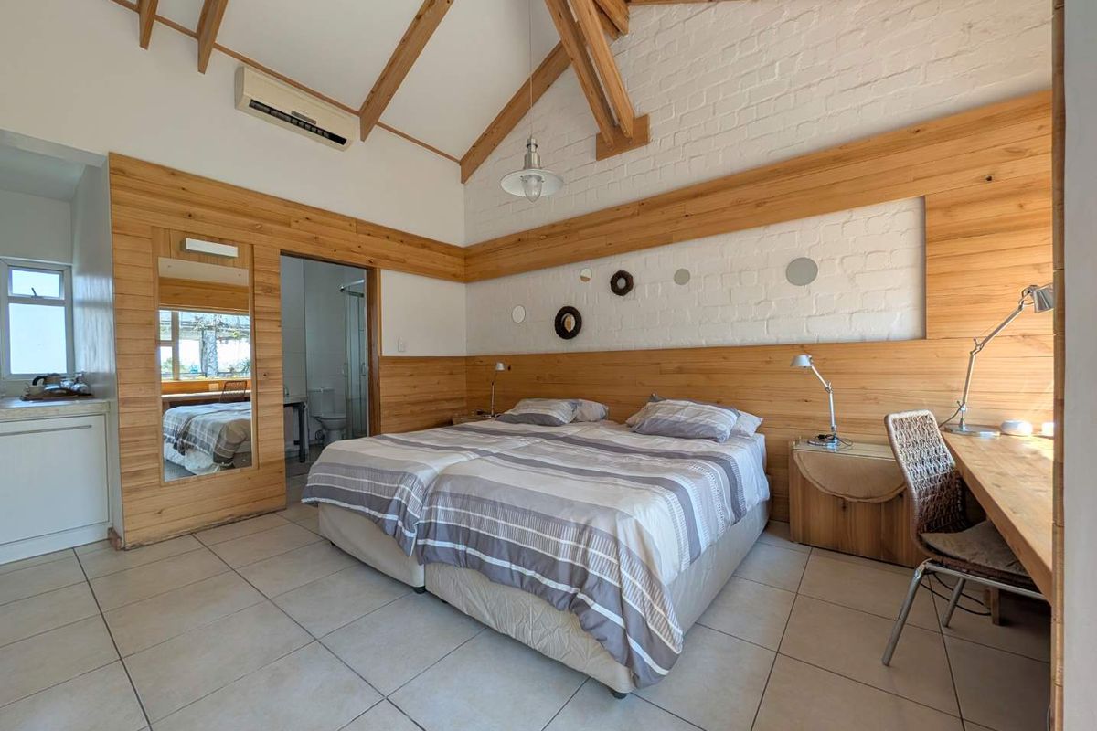 Spacious bedroom featuring exposed timber roof trusses, timber wall paneling, painted brickwork, tiled flooring, and an integrated desk area with en-suite bathroom access.