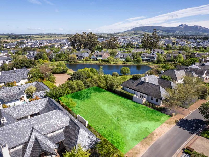 An aerial view of a vacant residential plot highlighted in green, situated within a well-established estate with surrounding homes and a nearby lake.