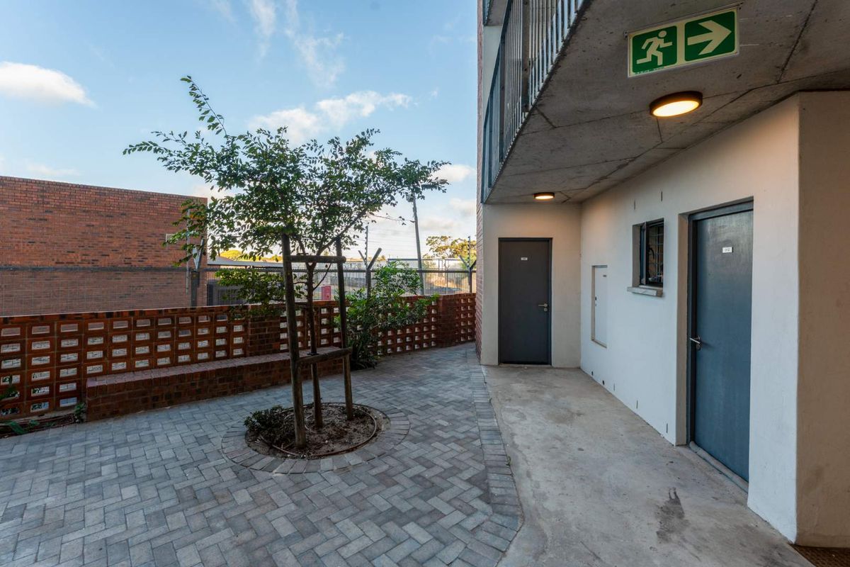 A paved outdoor courtyard area featuring brick boundary walls, a small tree, and access to apartment entry doors under a concrete overhang.