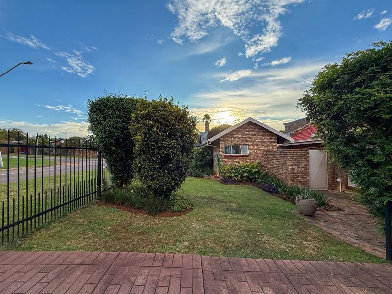 A well-maintained front exterior view of a brick house featuring a paved driveway, manicured lawn, and secure metal perimeter fencing.