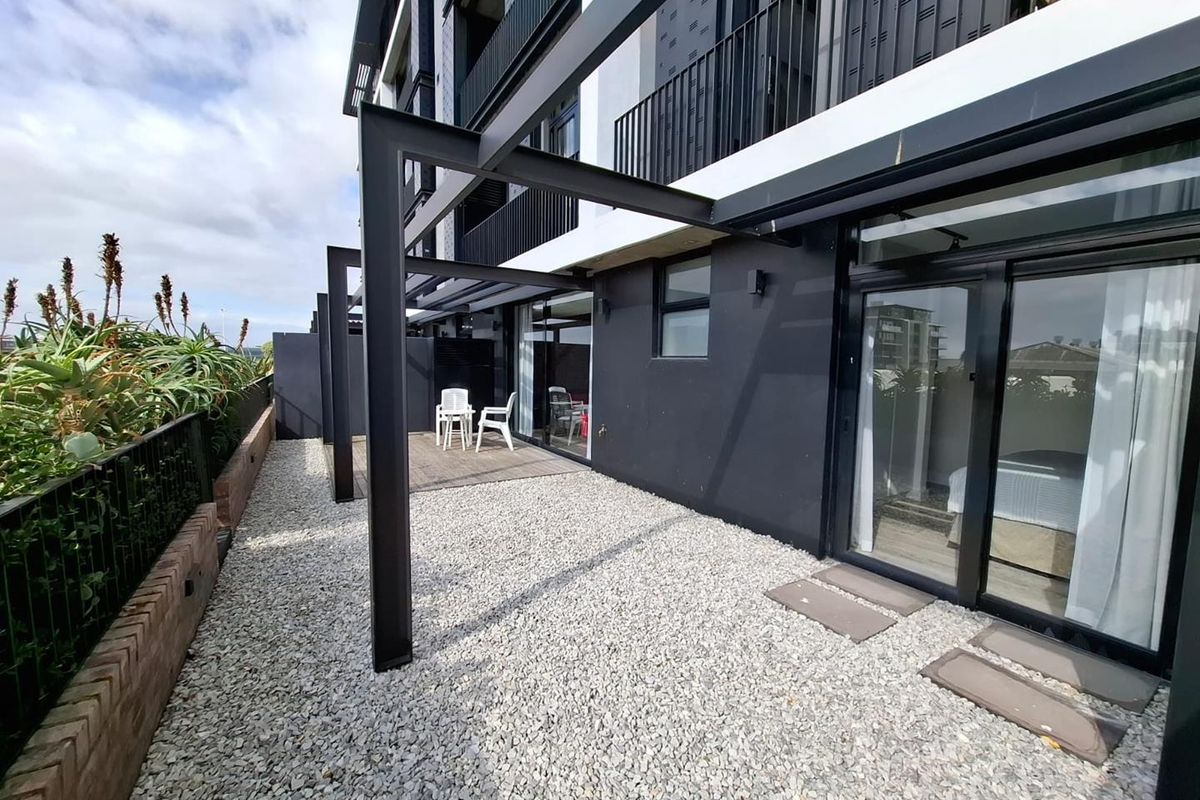 A modern ground floor apartment exterior featuring a gravel patio area, metal pergola structure, and sliding glass door access to the interior.