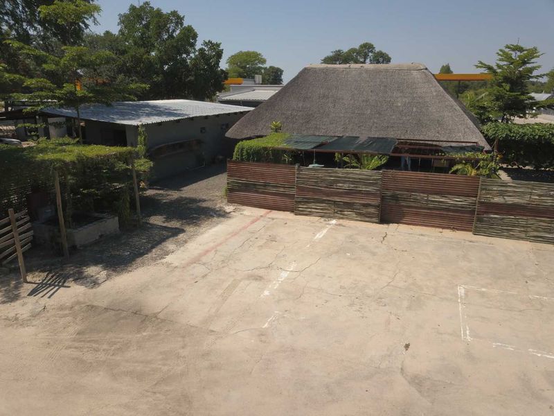 An aerial view of a rear yard area featuring a thatched roof structure, corrugated metal fencing, and a large concrete paved parking or utility space.