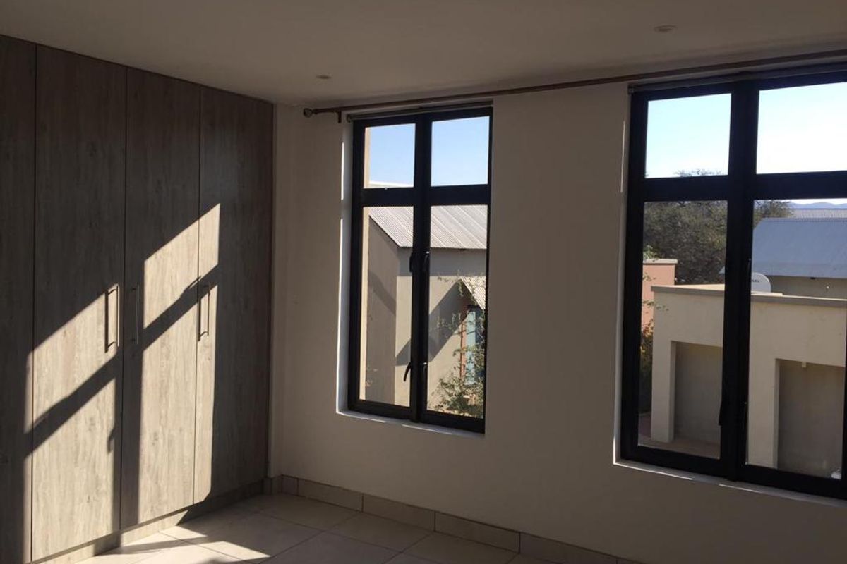 A bright bedroom featuring built-in wooden cabinetry, tiled flooring, and large windows with dark frames offering natural light and neighborhood views.