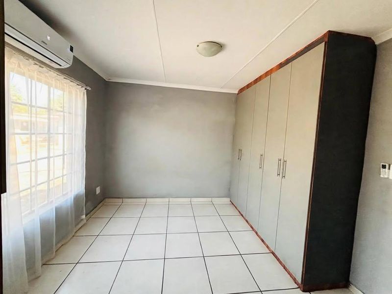 A clean, empty bedroom featuring neutral grey walls, white tiled flooring, built-in cupboards, and a wall-mounted air conditioning unit.