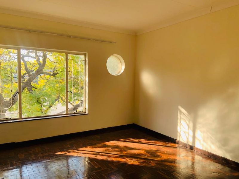 A bright, empty bedroom featuring classic herringbone parquet flooring, a unique circular window, and large windows providing natural light and garden views.