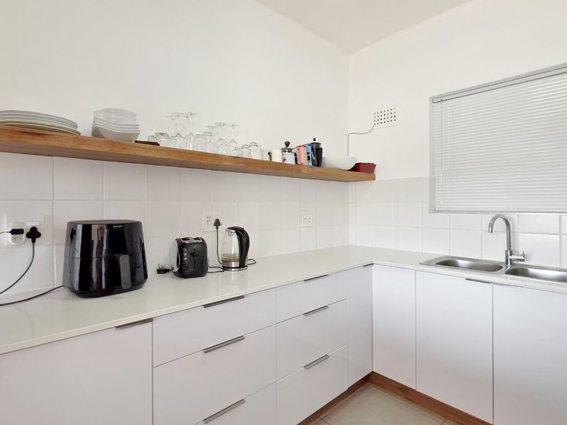 Modern kitchen featuring clean white cabinetry, stone countertops, a stainless steel sink, and a floating timber shelf with tiled splashback.