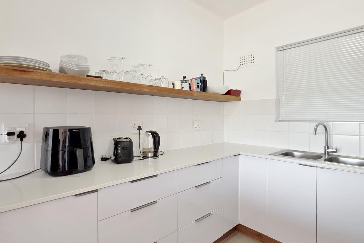 Modern kitchen featuring clean white cabinetry, stone countertops, a stainless steel sink, and a floating timber shelf with tiled splashback.