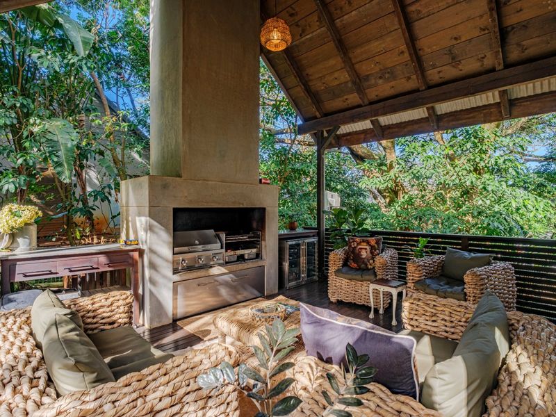 Covered outdoor patio featuring a built-in braai, timber decking, and exposed timber rafters, set within a lush, private garden environment.