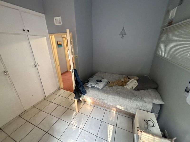 A compact bedroom featuring tiled flooring, built-in cupboards, and a window with blinds, offering a functional space with potential for cosmetic updates.