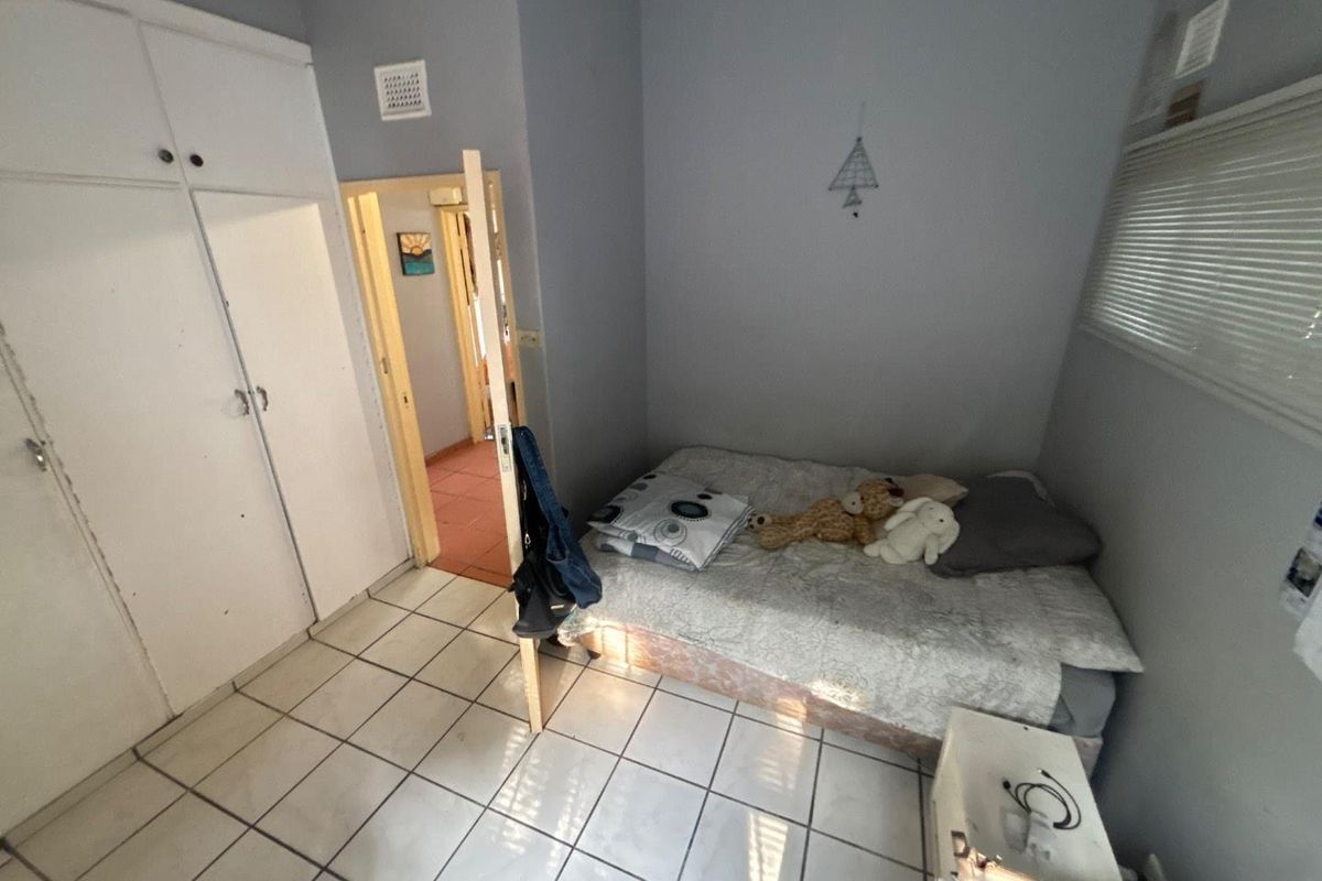 A compact bedroom featuring tiled flooring, built-in cupboards, and a window with blinds, offering a functional space with potential for cosmetic updates.