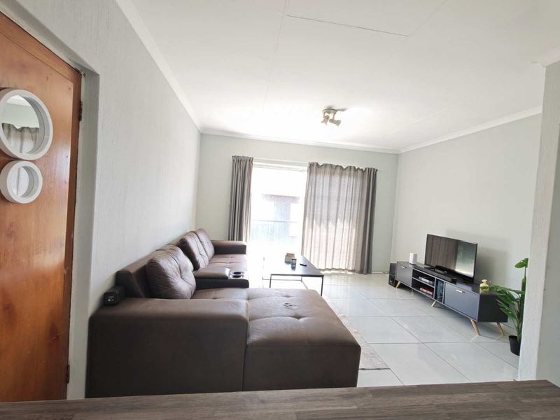 A bright, open-plan living room featuring neutral tiled flooring, coved ceilings, and a sliding glass door providing access to an outdoor balcony area.