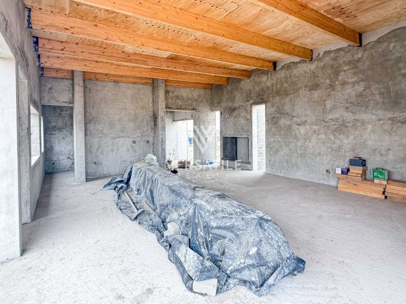 An expansive open-plan living area in a shell state, featuring exposed timber ceiling beams, raw cement walls, and a built-in fireplace.