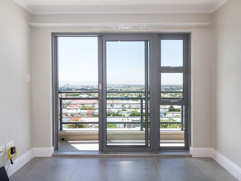 A bright, empty living area featuring modern grey floor tiles, neutral walls, and a large sliding glass door opening onto a balcony with city views.