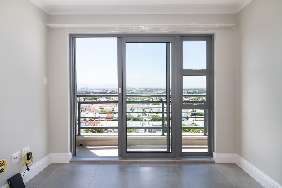 A bright, empty living area featuring modern grey floor tiles, neutral walls, and a large sliding glass door opening onto a balcony with city views.