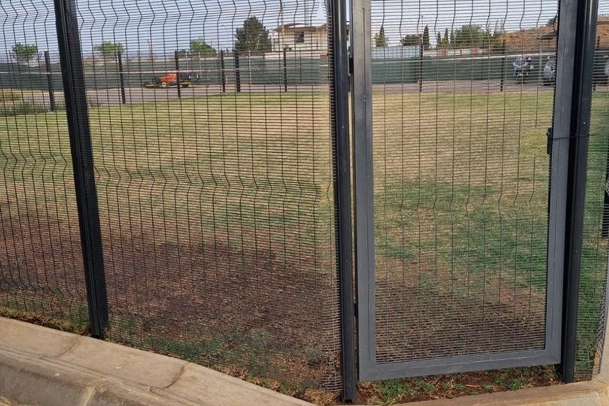 A secure perimeter featuring black steel mesh fencing and a matching pedestrian gate, bordering a paved walkway and an open grassed area.