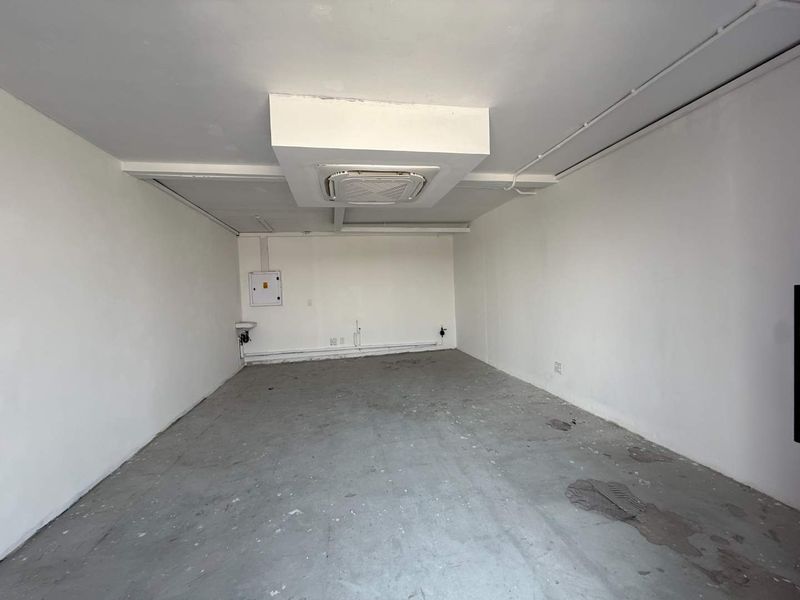 An empty, unfinished utility space featuring a ceiling-mounted air conditioning unit, a small wall-mounted basin, and exposed electrical infrastructure.