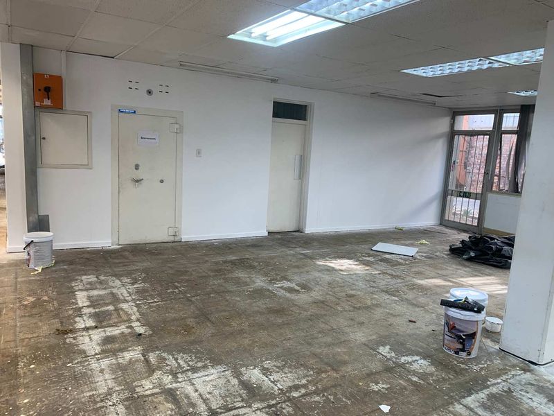 An empty industrial utility space featuring a security door, suspended ceiling with fluorescent lighting, and worn flooring requiring refurbishment.