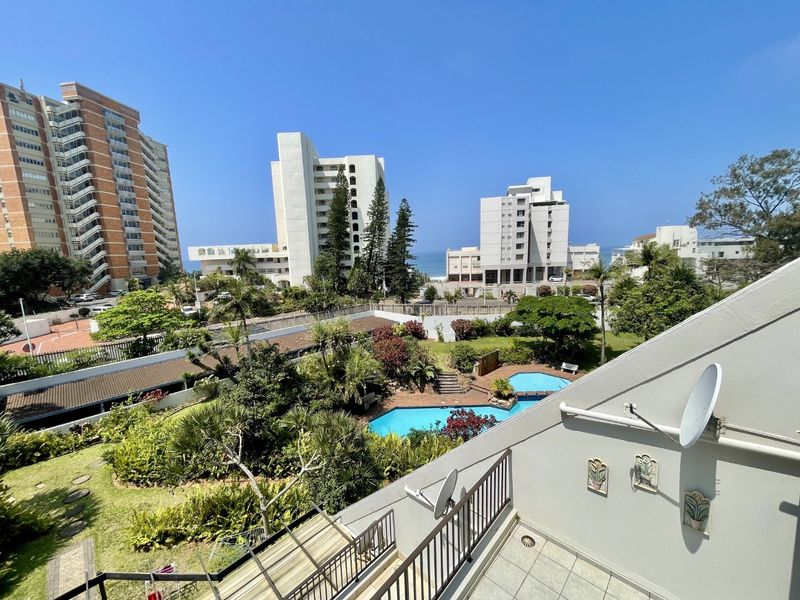 Elevated view overlooking a communal swimming pool, landscaped gardens, and surrounding apartment buildings with a glimpse of the ocean in the distance.
