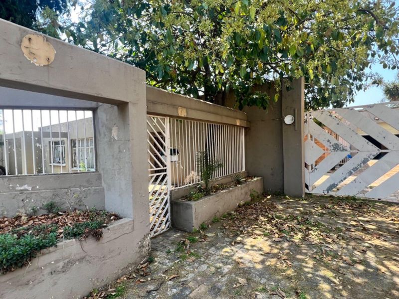 Front entrance area featuring a concrete boundary wall, metal security gate, and paved driveway, showing signs of wear and weathering.