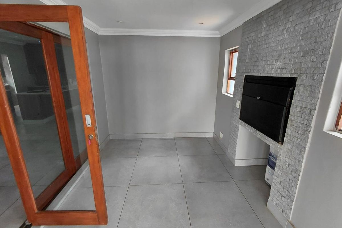 A bright living area featuring large grey floor tiles, a stone-clad fireplace, and wooden framed glass doors leading to an adjacent space.