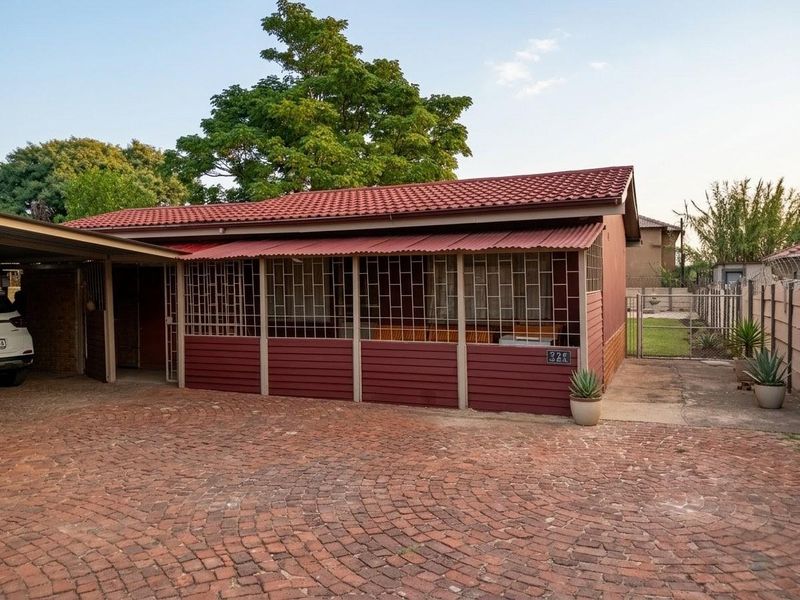 Front exterior view of a single-story house featuring a red metal roof, security bars on the patio, and a brick-paved driveway.