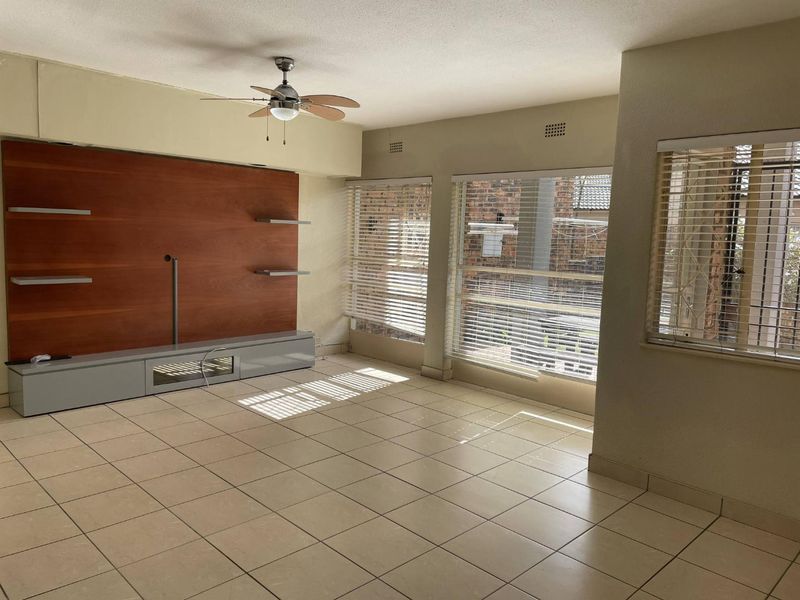 Spacious living room featuring neutral tiled flooring, a built-in media wall unit, ceiling fan, and large windows with fitted venetian blinds.