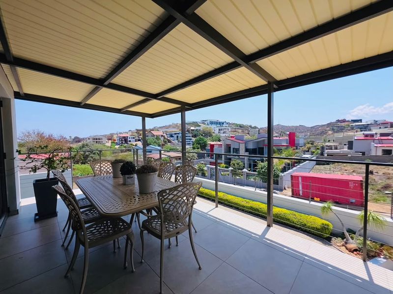 A spacious covered balcony featuring modern tiled flooring, a sleek metal roof structure, and a glass balustrade offering expansive neighbourhood views.