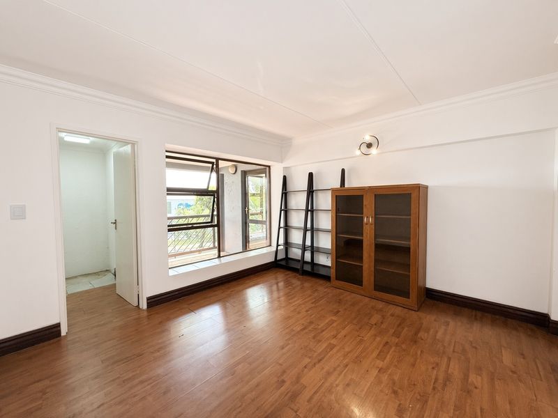 A bright bedroom featuring laminate flooring, white walls with decorative cornicing, and large aluminium windows allowing for natural light.