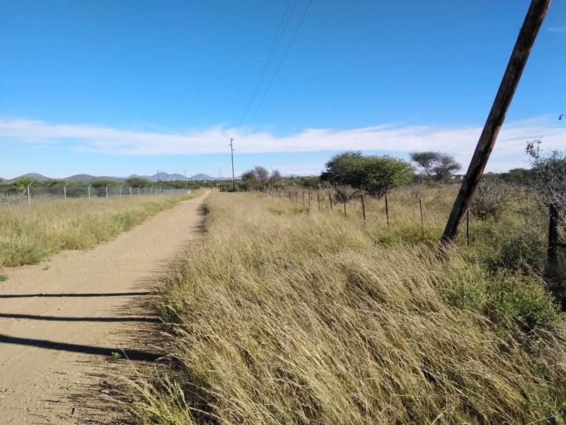 A view of a vacant residential plot featuring a dirt access road, wire perimeter fencing, and utility infrastructure under a clear blue sky.