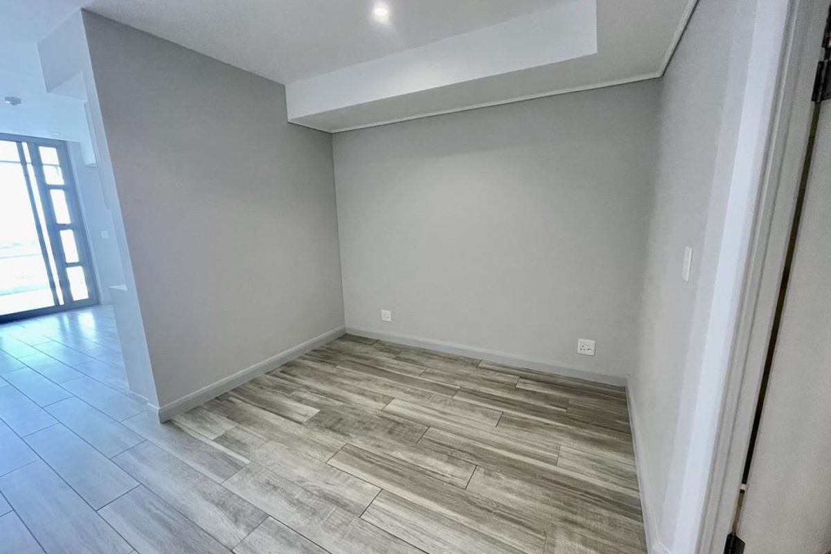 A bright, empty room featuring modern wood-look tile flooring, neutral grey walls, and a recessed ceiling detail, suitable for a bedroom or home office.