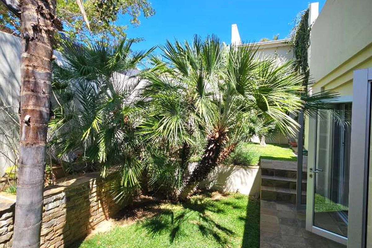 A private rear garden featuring a paved patio area, lush lawn, mature palm trees, and stone retaining walls, accessible via glass sliding doors.