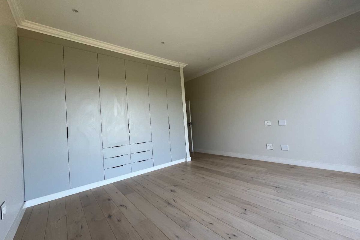 A bright, empty bedroom featuring modern light-toned engineered wood flooring, neutral walls, and extensive floor-to-ceiling built-in cupboards with integrated drawers.