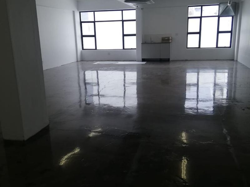 An open-plan office space featuring polished concrete floors, exposed industrial ceiling ducting, and a small kitchenette area with large windows providing natural light.