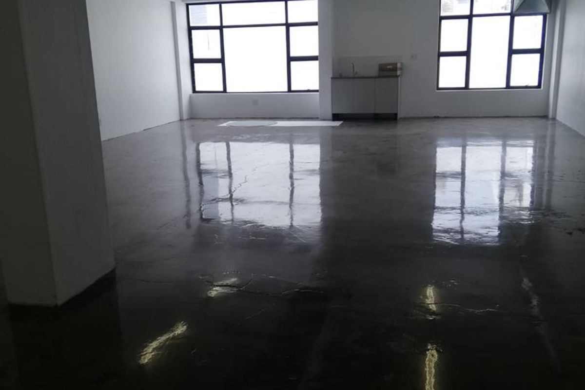 An open-plan office space featuring polished concrete floors, exposed industrial ceiling ducting, and a small kitchenette area with large windows providing natural light.
