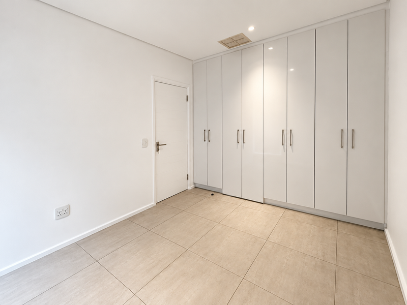 A bright, empty bedroom featuring modern floor-to-ceiling built-in cupboards, neutral tiled flooring, and recessed ceiling lighting.