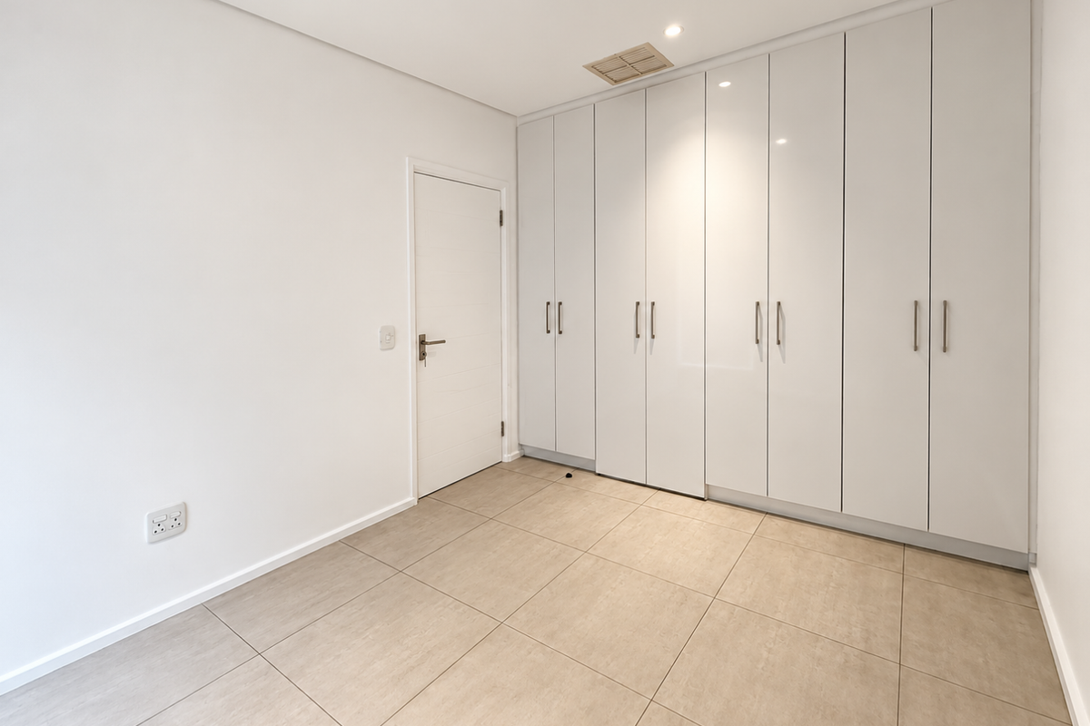 A bright, empty bedroom featuring modern floor-to-ceiling built-in cupboards, neutral tiled flooring, and recessed ceiling lighting.