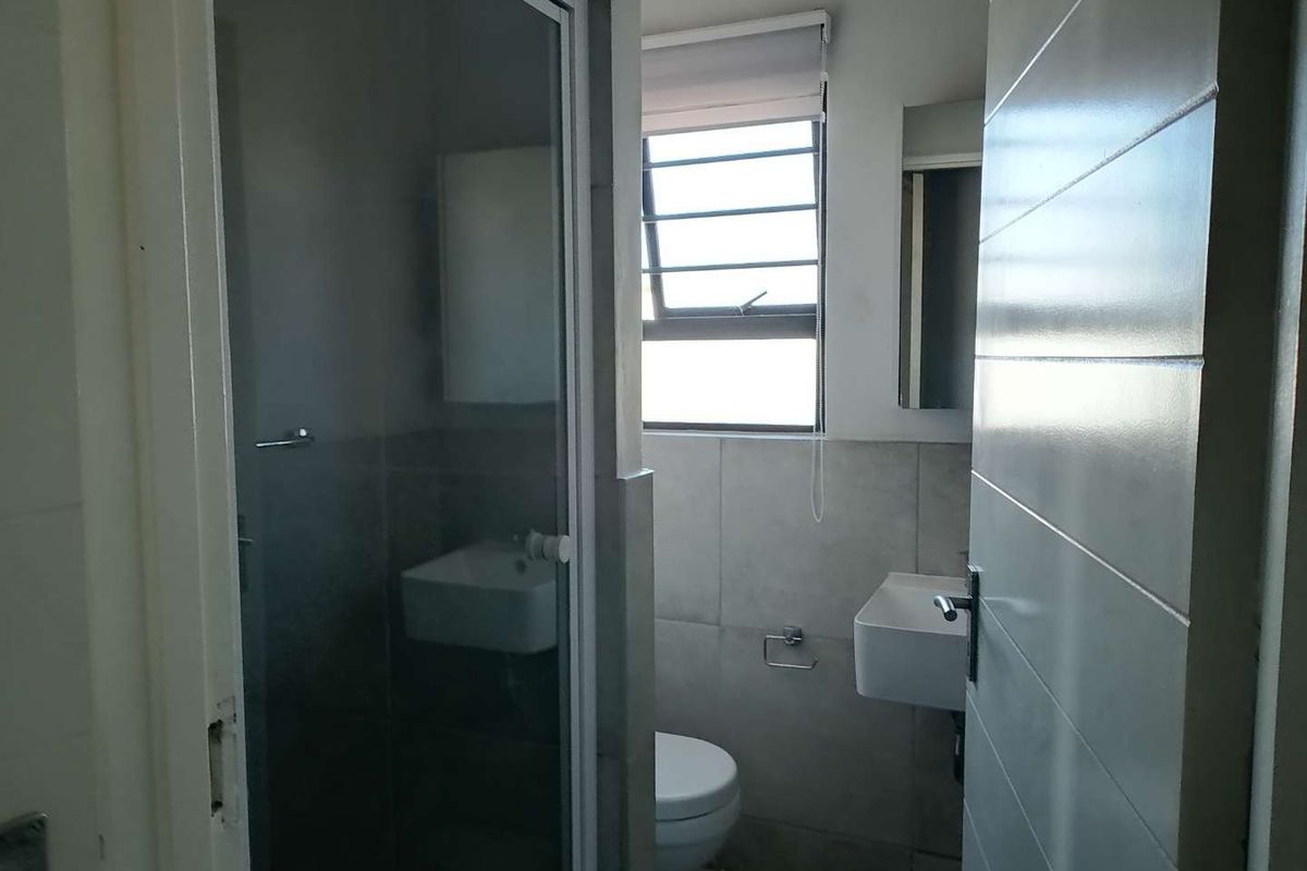 A functional bathroom featuring a glass-enclosed shower, wall-mounted basin, and toilet, finished with neutral wall tiles and a window for natural light.