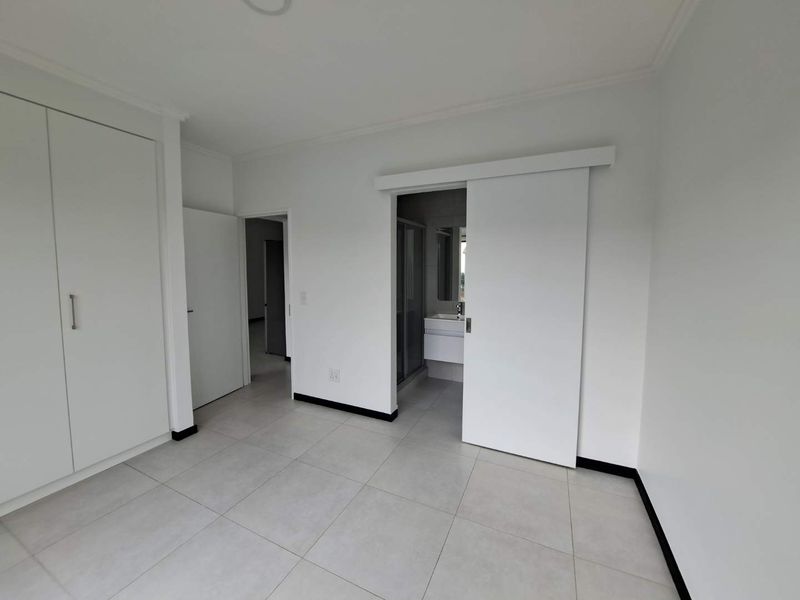 A clean, modern bedroom featuring neutral tiled flooring, built-in cupboards, and a sliding door providing access to an en-suite bathroom.