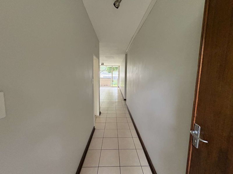 A clean, empty hallway featuring neutral tiled flooring and simple coving, leading towards an open-plan living area with natural light.