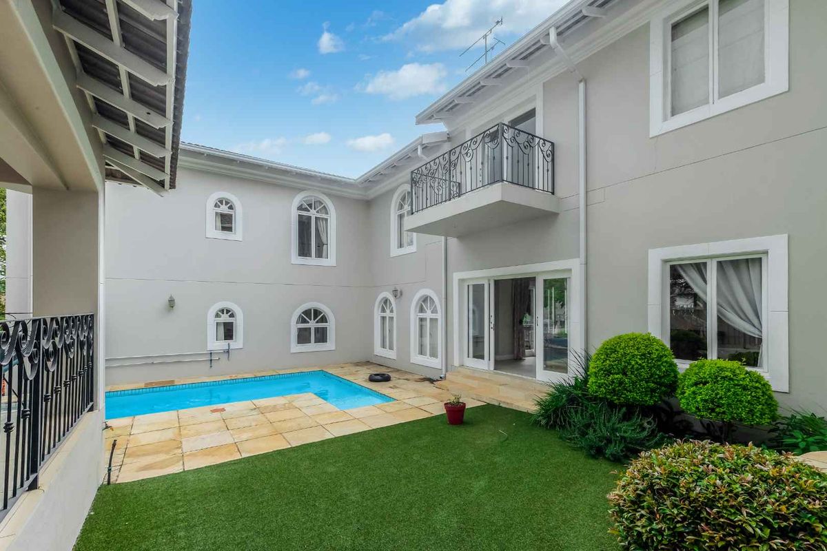 A well-maintained rear exterior view featuring a swimming pool, paved patio area, artificial lawn, and a balcony with decorative wrought iron railings.