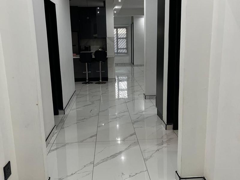 A bright, modern hallway featuring polished marble-look floor tiles, clean white walls, and recessed lighting, leading towards an open-plan kitchen area.