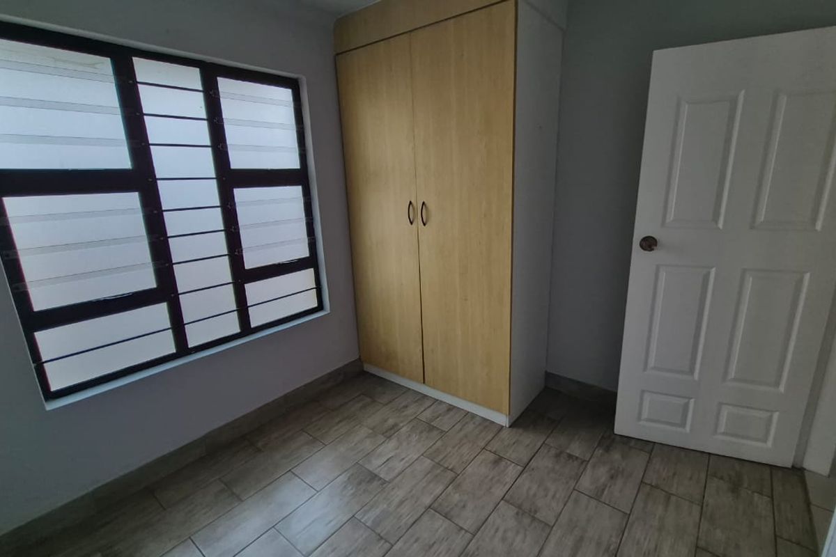 A clean, empty bedroom featuring wood-look tiled flooring, a built-in wardrobe, and a window fitted with security bars.