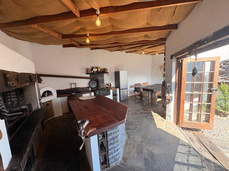 Covered outdoor entertainment area featuring a built-in pizza oven, stone bar, fireplace, and rustic timber ceiling, opening onto a garden space.