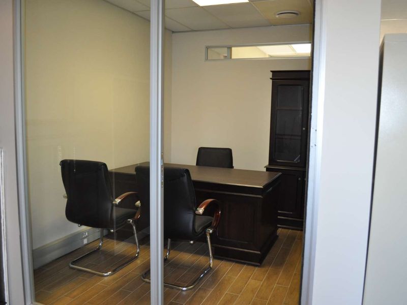 A professional office space featuring a glass partition wall, wood-look tile flooring, and a suspended ceiling with recessed lighting.