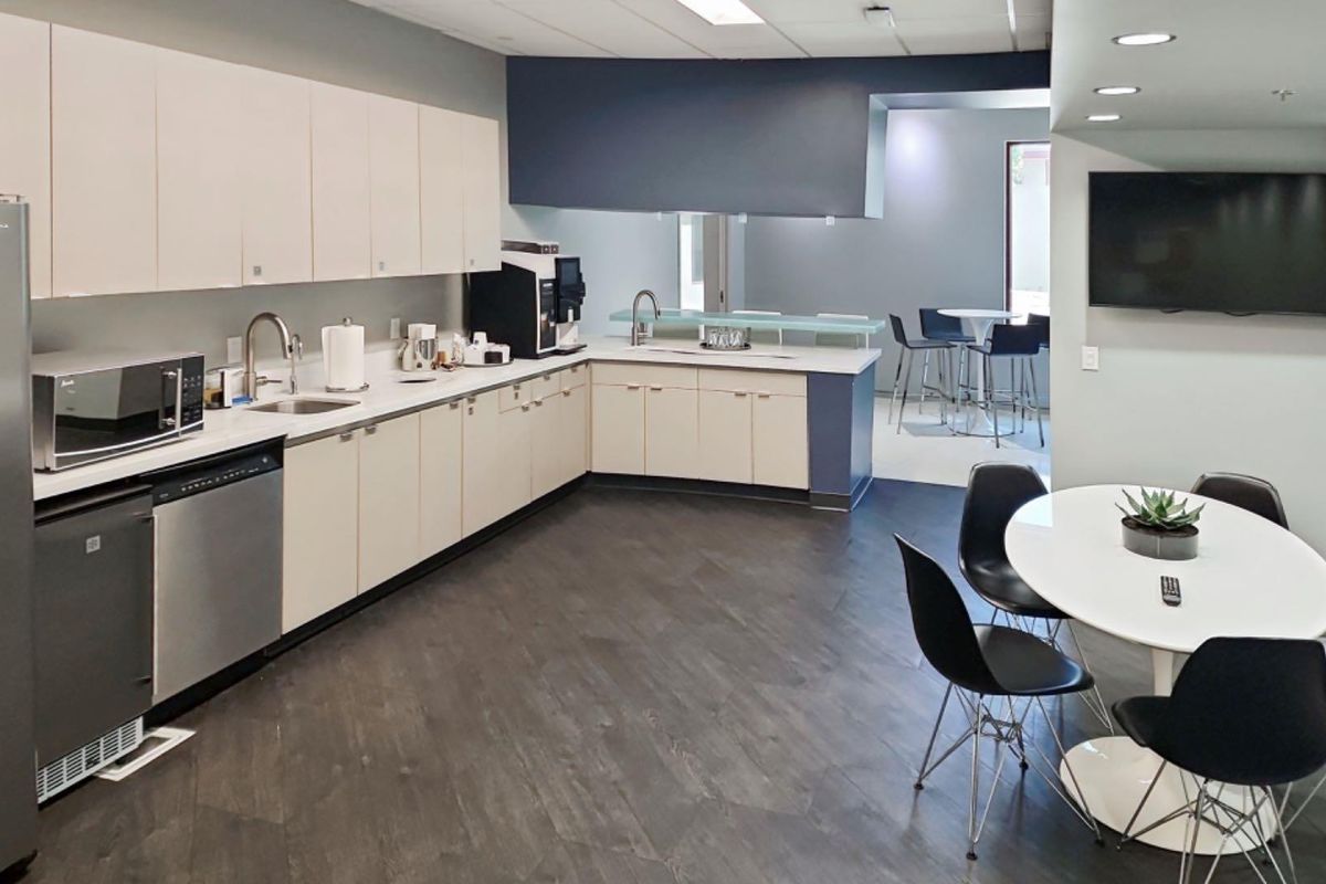 A modern office kitchenette featuring clean white cabinetry, laminate countertops, stainless steel sink, integrated dishwasher, and durable dark wood-look flooring.