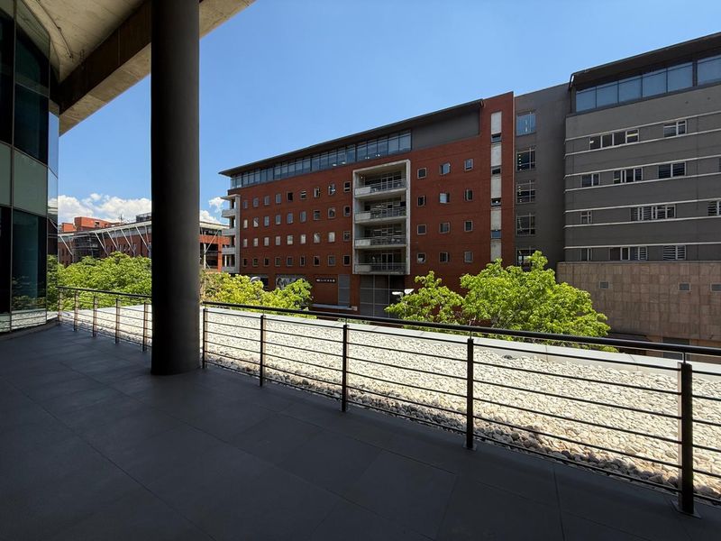 A spacious covered balcony featuring tiled flooring, a stainless steel railing, and views overlooking the surrounding office complex and urban landscape.