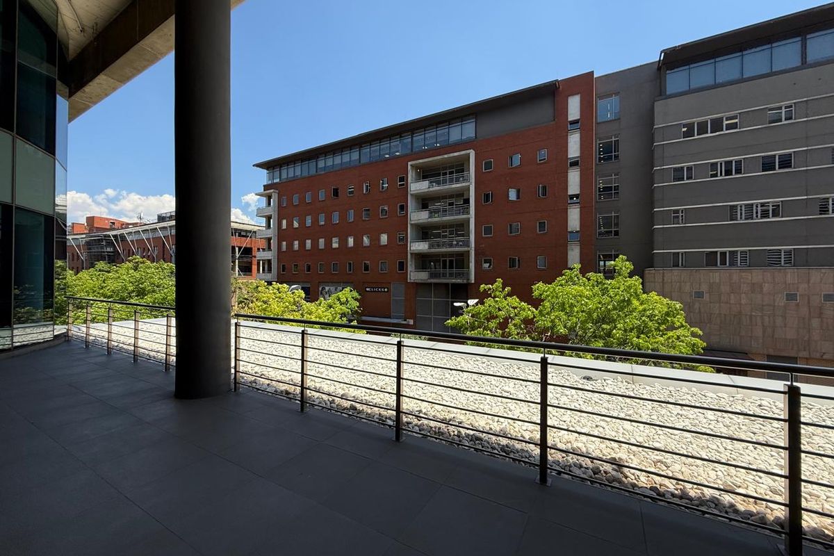 A spacious covered balcony featuring tiled flooring, a stainless steel railing, and views overlooking the surrounding office complex and urban landscape.